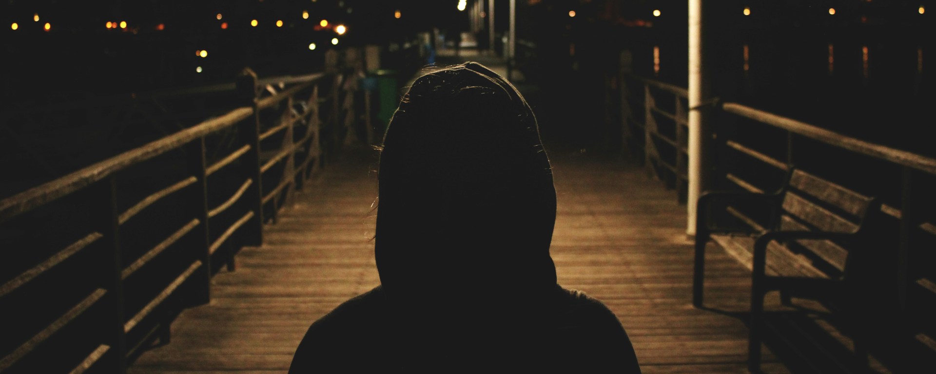 Uma pessoa de costas voltadas para a imagem, vestida com um capaz, numa noite escura onde apenas se vê um poste de luz mais adiante.